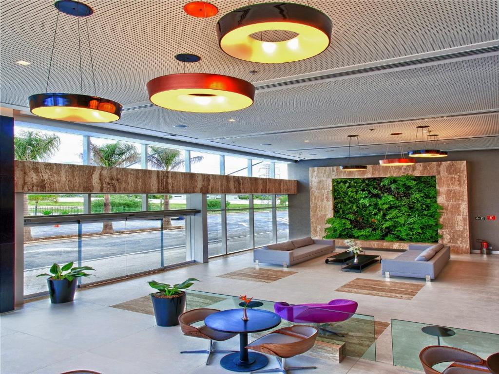 Área da recepção do Golden Tulip São José dos Campos, um dos hotéis indicados na cidade, com sofás em frente a uma parede com jardim suspenso, poltronas coloridas e mesinha, além de lustres redondos no pé direito alto