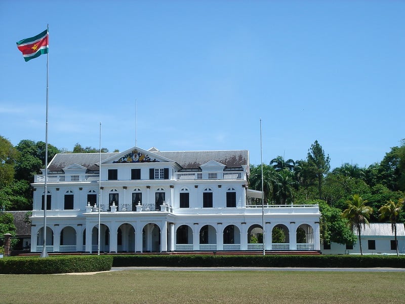 Palácio branco com bastante janelas em frente de um gramado verde e com árvores ao fundo durante o dia, ilustrando post chip celular Suriname.