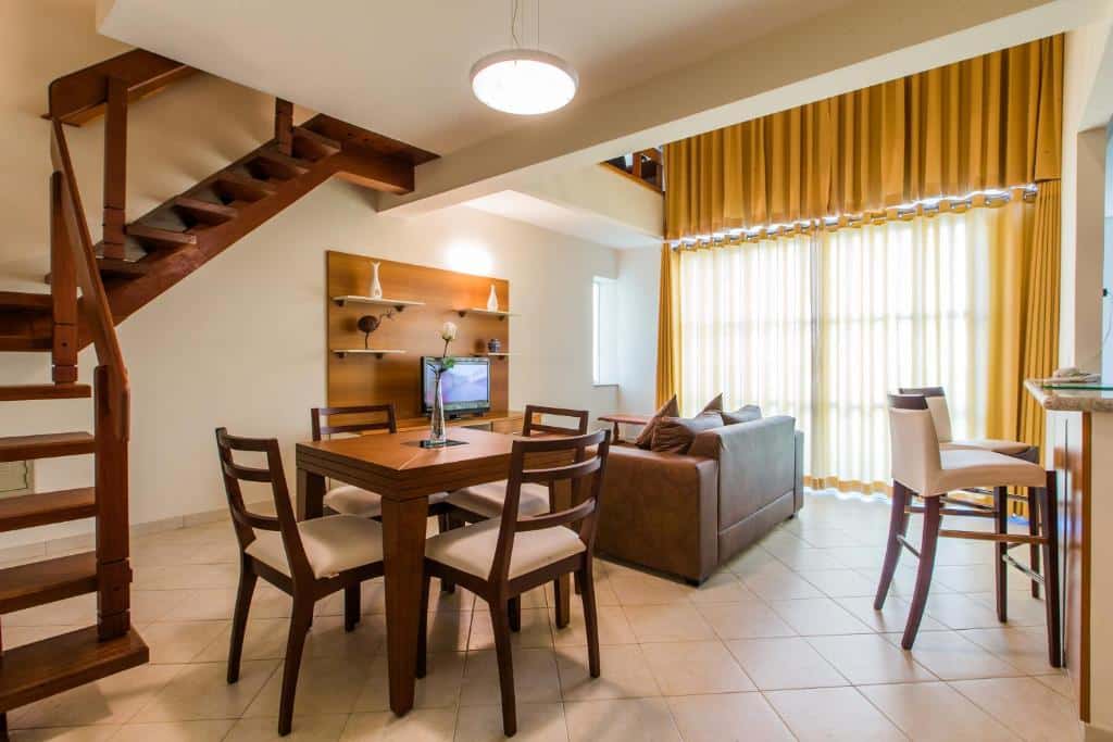 Sala de opção de apartamento duplex no Hotel City Hall, em São José dos Campos, com escada de madeira, mesa quadrada com quatro lugares, banquetas na bancada da cozinha, sofá virado para a TV, e cortina