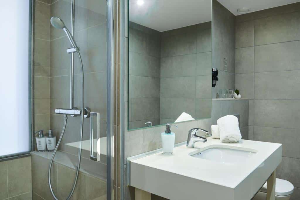 Banheiro do Hotel Omnium. Do lado esquerdo da foto há um box com chuveiro e amenidades de banho. Ao lado, uma pia branca tem toalhas e sabonete em cima e um espelho está na parede acima. Ao fundo há uma privada branca.
