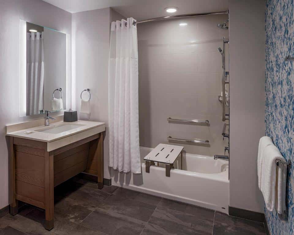 Banheiro adaptado do Hyatt House Tampa Downtown com uma banheira com barras de apoio e um banco para se sentar, pia mais baixa e local espaçoso para uma cadeira de rodas