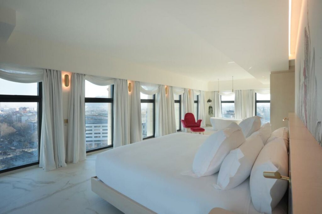 Quarto do Iberostar Selection Paseo de Gracia, uma das recomendações de hotéis no centro de Barcelona. A cama com lençol e travesseiros brancos está de frente para uma parede repleta de janelas com vista para a cidade. Há luminárias dos dois lados da cebceira da cama. Ao fundo do quarto há uma poltrona vermelha.