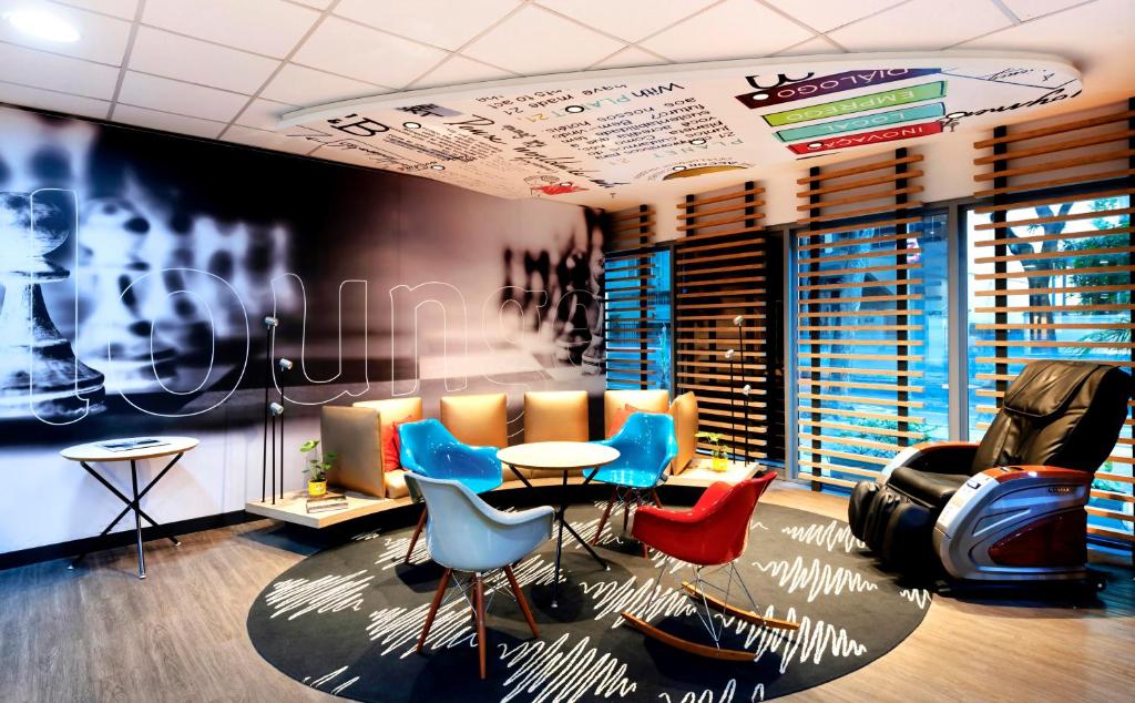 Imagem do lounge compartilhado do hotel com cadeira de massagem, uma mesa no centro com cadeiras coloridas, tapete preto com detalhes brancos, teto colorido, e uma parede escrito lounge.