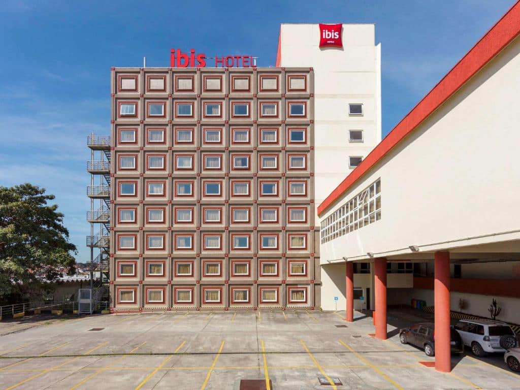 Área externa do hotel Ibis São José dos Campos Dutra, com grande estacionamento, de vagas ao ar livre e cobertas. Ao fundo aparece o prédio com os quartos do hotel