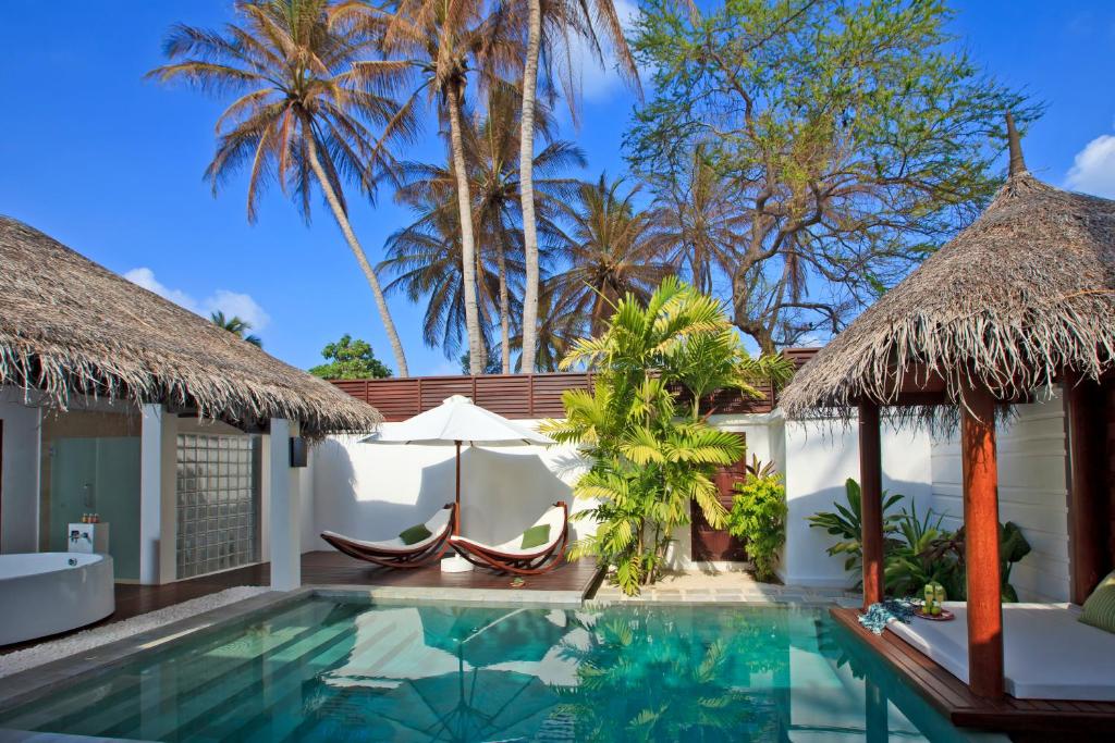 vista da área externa de um quarto com piscina e bangalô para deitar, duas espreguiçadeiras estilosas e entrada para o quarto do Velassaru Maldives com palmeiras do lado de fora