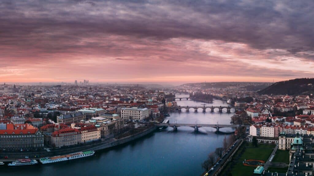 Vista aérea de Praga, é possível avistar três pontes cortando o rio principal da cidade com muitos prédios antigos e algumas embarcações estacionadas