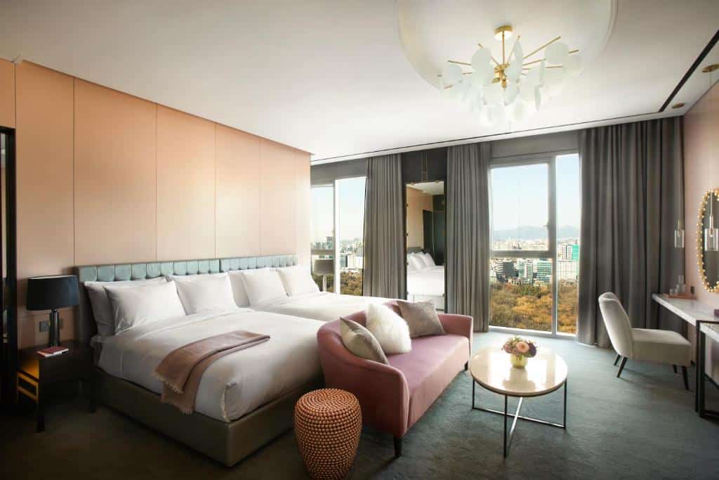 Quarto do L7 Gangnam by LOTTE com uma janela panorâmica com cortinas, um lustre, um carpete cinza, duas camas de casal, um sofá com almofadas e uma bancada com uma cadeira com um espelho na frente