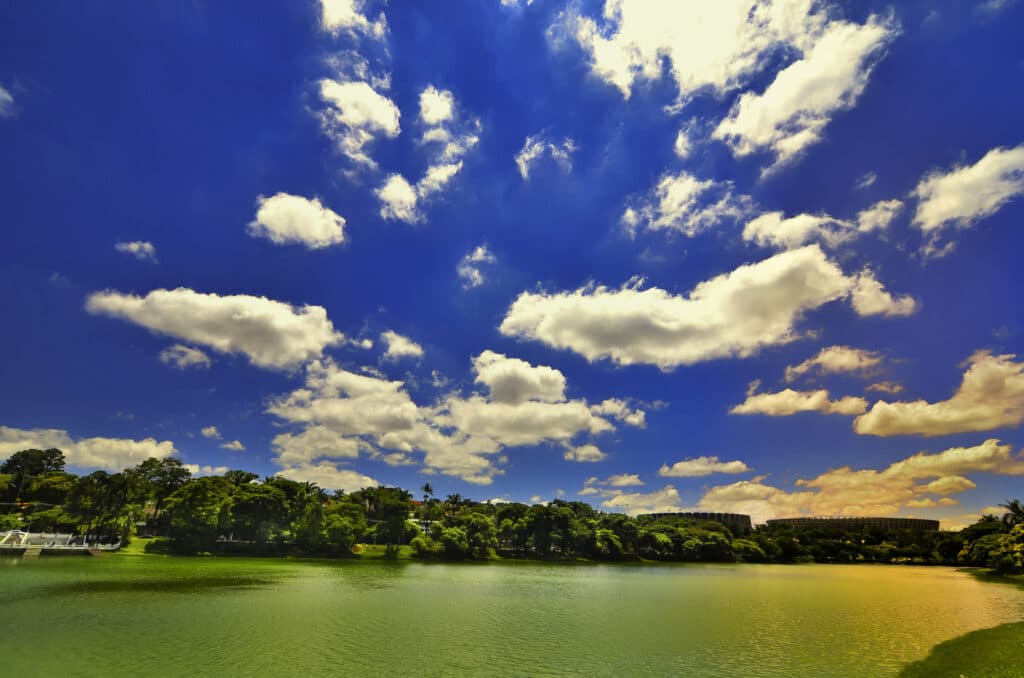 vista da Lagoa da Pampulha, em Belo Horizonte, com águas esverdeadas e muita vegetação ao redor, o céu é azul com muitas nuvens
