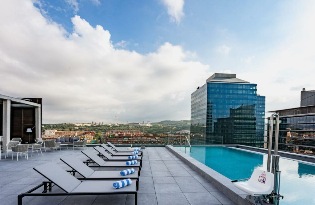 Piscina do Leonardo Royal Hotel Barcelona Fira, uma das recomendações de hotéis baratos em Barcelona. Espreguiçadeiras com toalhas em cima estão por toda a extensão da piscina que fica na cobertura. Há um elevador de piscina na borda. Ao fundo é possível ver a cidade de Barcelona.