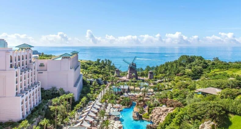 Vista aérea do Lotte Hotel Jeju, com piscina, muitas árvores cercando a propriedade que está de frente para o mar, e conta com alguns moinhos dentro da propriedade, para representar hotéis na Coreia do Sul