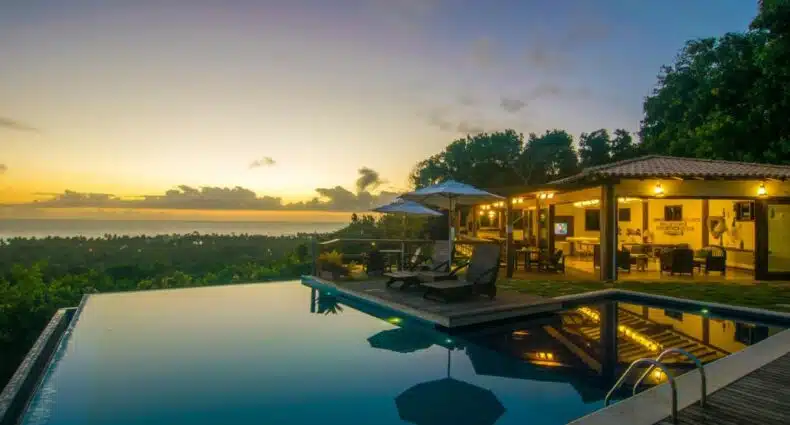 Piscina da Manacá Pousada Parque, uma das recomendações de pousadas em Arraial D'Ajuda. Duas espreguiçadeiras na borda da piscina têm um guarda-sol no meio. Ao fundo há uma área coberta de recreação com mesas e cadeiras.