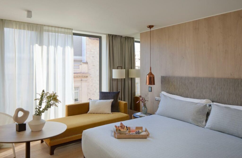 Quarto do hotel ME Barcelona, uma das recomendações de hotéis no centro de Barcelona. Há uma bandeja de café da manhã em cima da cama com lençol e travesseiros cinza e brancos. Há uma mesinha de cabeceira com um abajur pendurado dos dois lados da cama.
Um divã amarelo com almofadas está encostado em uma janela coberta por cortinas cinza e brancas. Há uma mesa com uma planta no canto esquerdo da imagem, e um abajur atrás do divã.