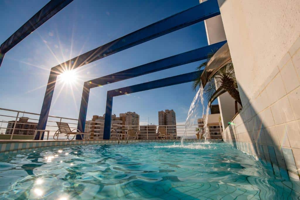 Piscina no terraço do Mondrian Suite Hotel, em São José dos Campos, em dia de sol forte e céu azul sem nuvens. Há duas cascatas sobre a piscina, e cadeiras para tomar sol ao redor