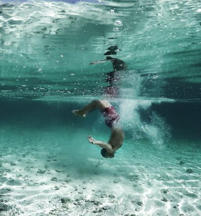 Homem de ponta cabeça embaixo d'água. Ele veste uma bermuda vermelha, e a água ao redor é transparente. - Foto: Moon via Unsplash