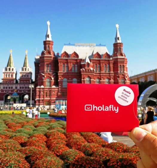 Uma mão branca segurando um cartão de chip vermelho escrito Holafly no meio e, de fundo, o Museu Histórico do Estado da Rússia com um jardim e prédio histórico vermelho com torres. Representa o post Holafly é confiável