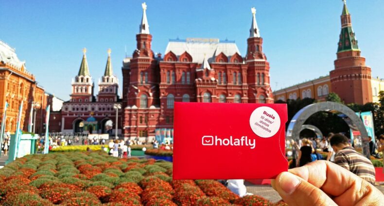 Uma mão branca segurando um cartão de chip vermelho escrito Holafly no meio e, de fundo, o Museu Histórico do Estado da Rússia com um jardim e prédio histórico vermelho com torres. Representa o post Holafly é confiável