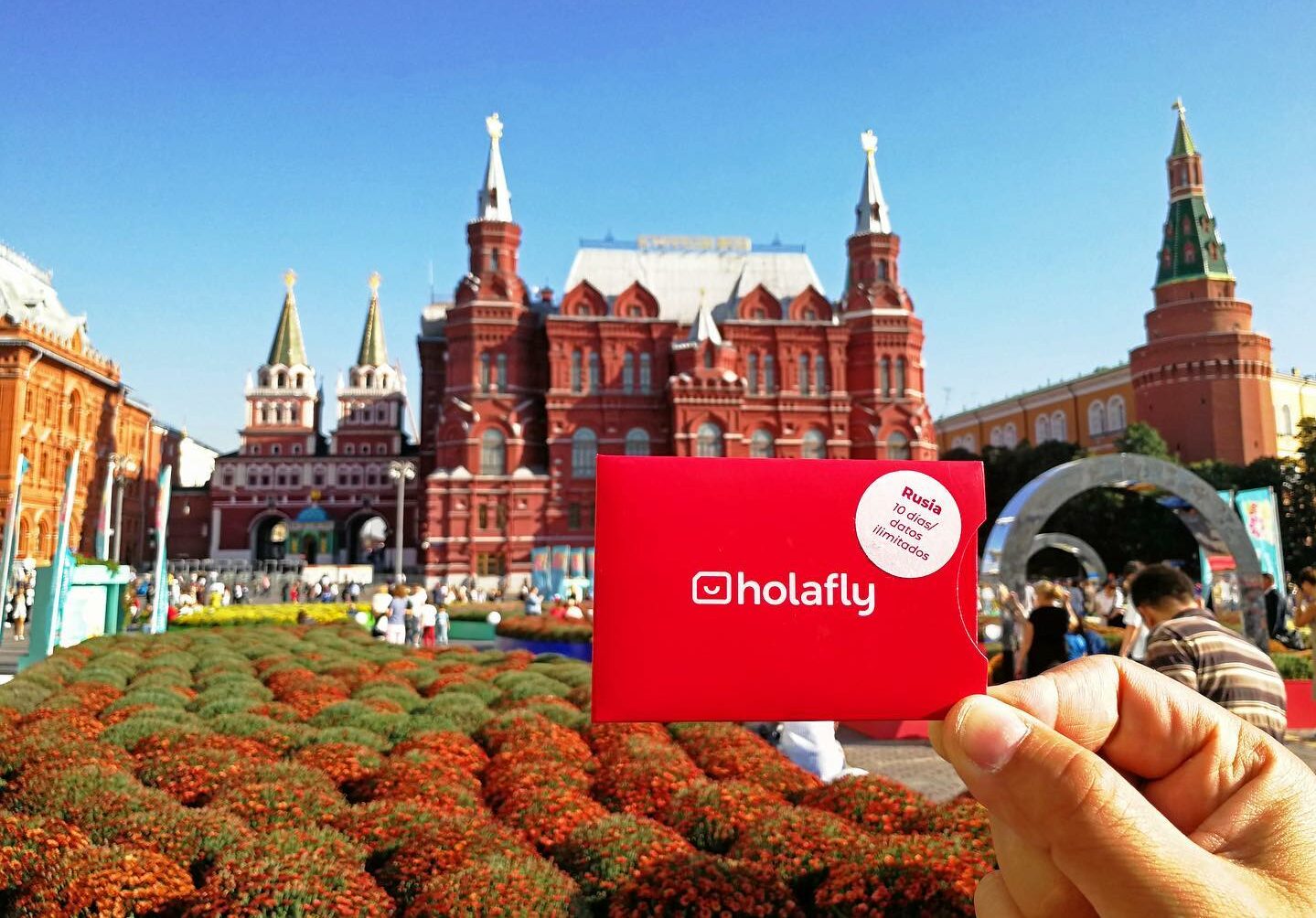 Uma mão branca segurando um cartão de chip vermelho escrito Holafly no meio e, de fundo, o Museu Histórico do Estado da Rússia com um jardim e prédio histórico vermelho com torres. Representa o post Holafly é confiável