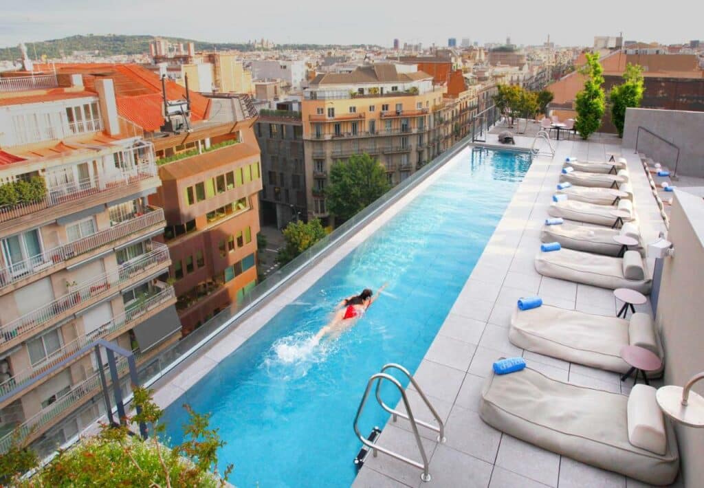 Piscina do Ohla Eixample, uma das recomendações de onde ficar em Barcelona. Há algumas almofadas de piscina do lado direito, e a piscina se estende por toda a beira do terraço. é possível ver grande parte da cidade.