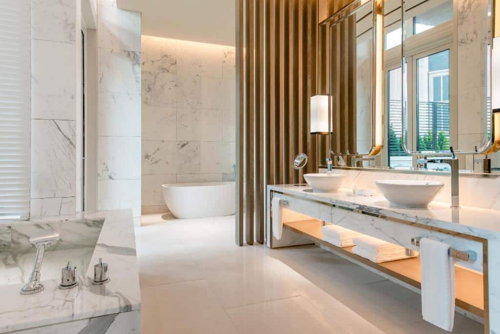 Banheiro do Paradise City com uma banheira oval, uma pia de mármore com duas cubas, espelhos, e algumas toalhas brancas pelo ambiente