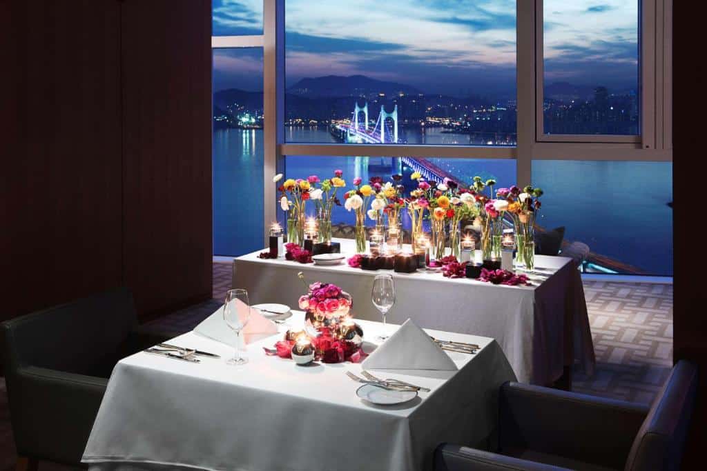 Uma mesa de jantar para duas pessoas no Park Hyatt Busan ao lado de uma janela ampla com vista para uma ponte iluminada, para representar hotéis na Coreia do Sul