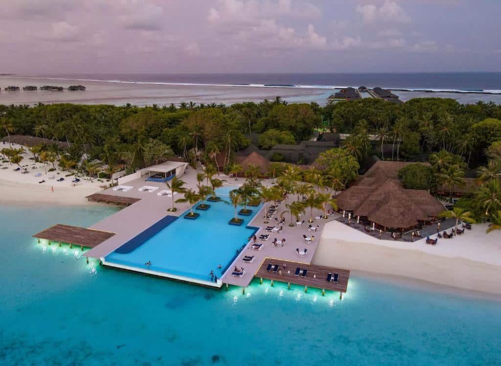 vista de cima do mar cristalino e da piscina do Paradise Island Resort & Spa, um dos resorts nas Maldivas, com alguns bangalôs e várias cadeiras em volta da piscina e na praia de areias brancas