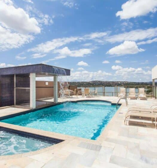piscina no terraço do Ramada Hotel & Suites Lagoa Santa By Wyndham, um dos hotéis perto do Aeroporto de Confins, com fonte de água e espreguiçadeiras, o céu é azul com nuvens e há vista para a lagoa