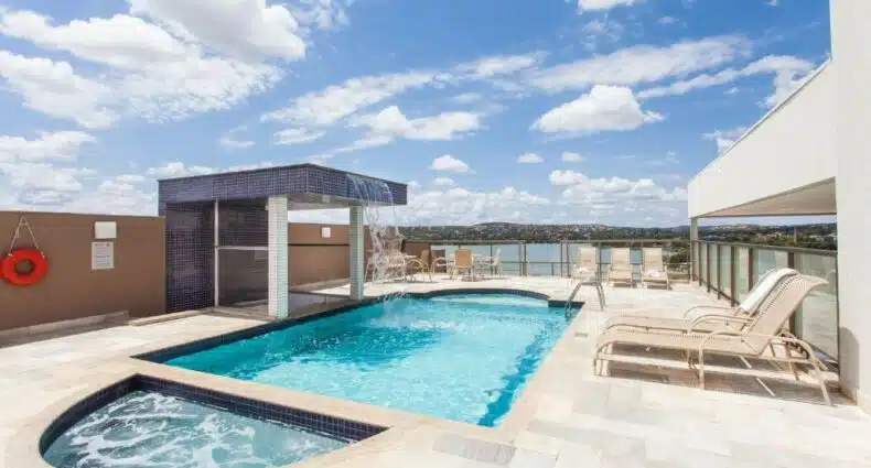 piscina no terraço do Ramada Hotel & Suites Lagoa Santa By Wyndham, um dos hotéis perto do Aeroporto de Confins, com fonte de água e espreguiçadeiras, o céu é azul com nuvens e há vista para a lagoa