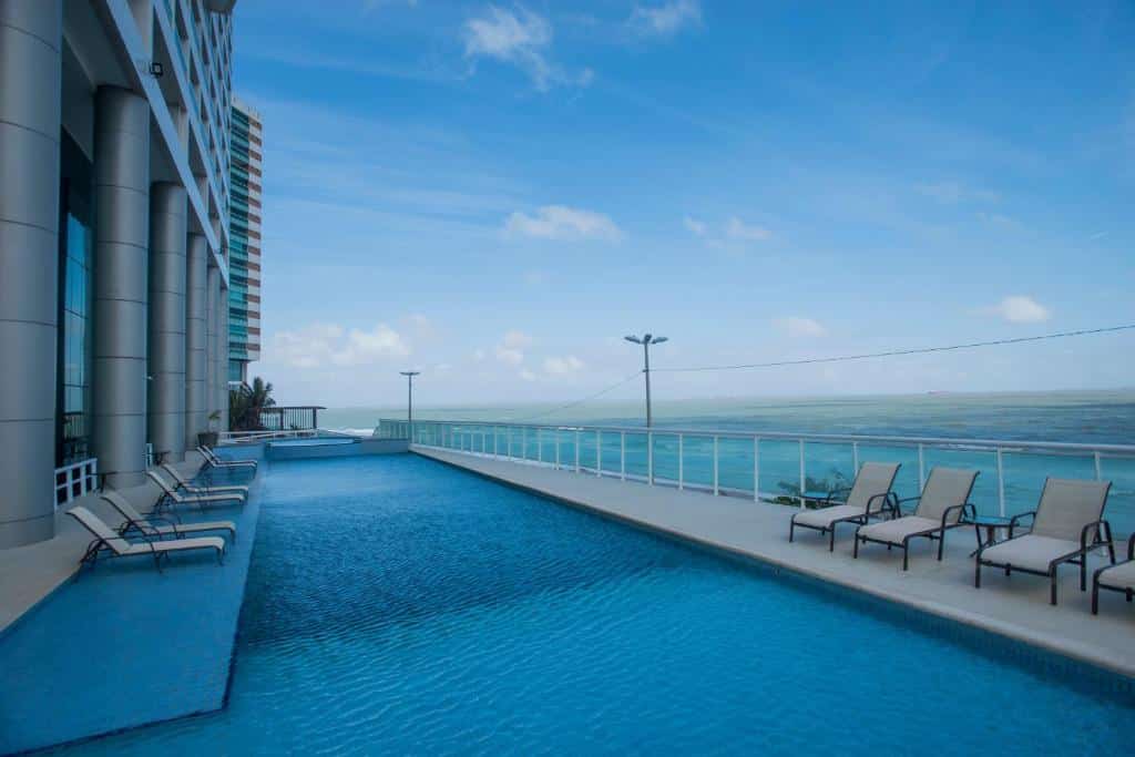 Vista da piscina do Hotel Luzeiros São Luis com cadeiras do lado direito e esquerdo e a piscina no meio e ao fundo vista para o mar. Representa São Luis do Maranhão.