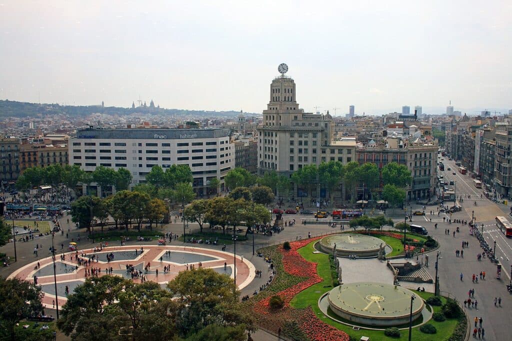 Foto da Plaça Catalunya vista de cima. Há construções ao redor e pessoas passeando pelas ruas. - Foto: via Wikimedia
