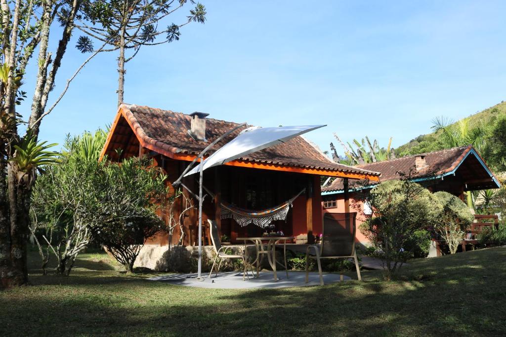 Casa com rede na varanda, mesa com duas cadeiras e guarda-sol em frente e árvores em volta durante o dia, ilustrando post pousadas em Visconde de Mauá.