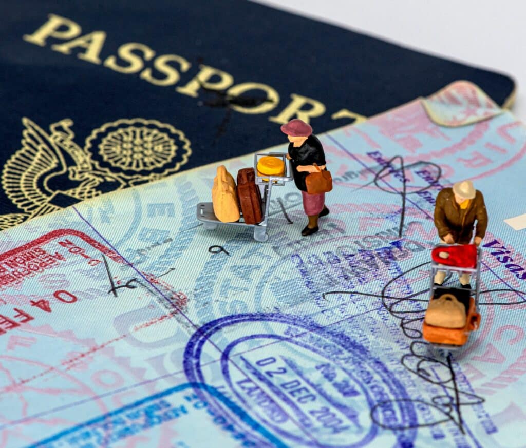 Duas figuras, uma de mulher e uma de homem, carregando carrinhos de aeroporto cheios de malas, posicionados sobre a página de um passaporte cheio de carimbos
