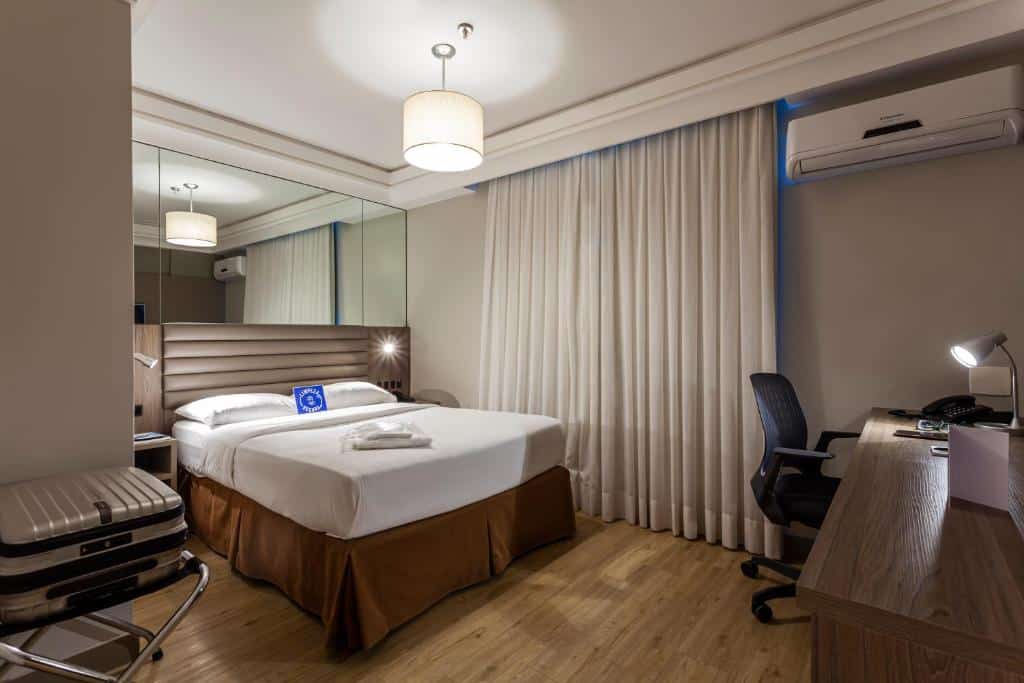 quarto do Blue Tree Premium Florianópolis com cama de casal e espelho atrás, com janela com cortina clara, mesa e cadeira e luminária de teto