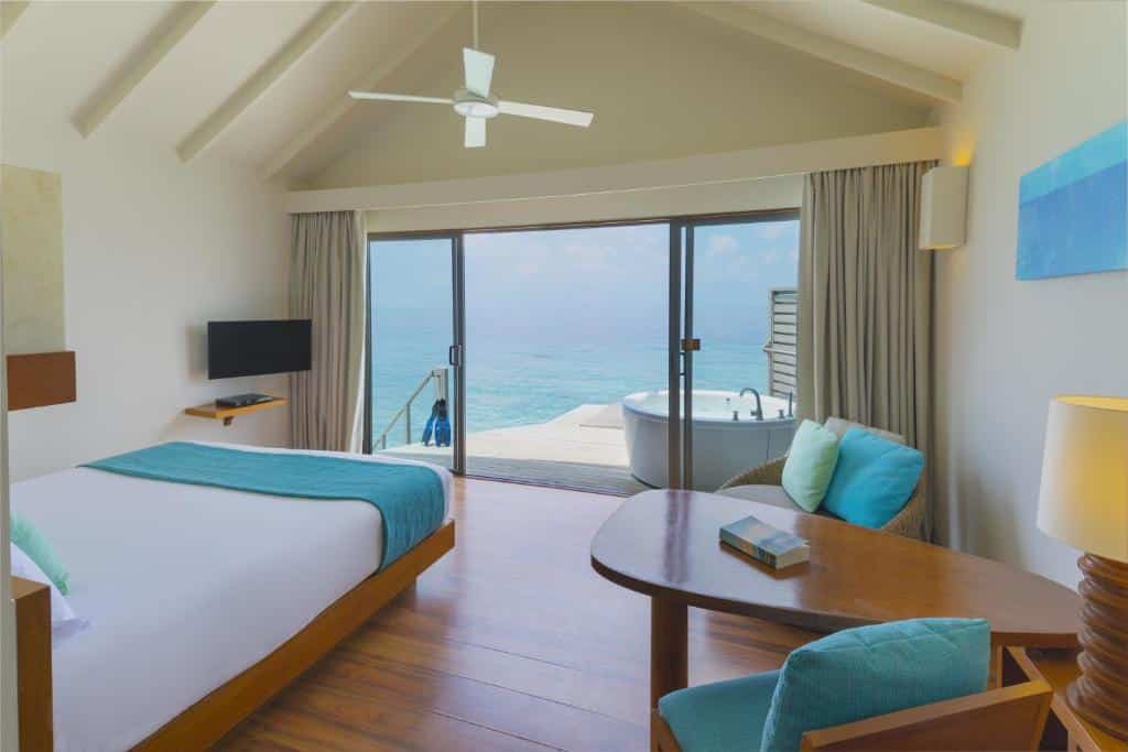 quarto do Centara Ras Fushi Resort & Spa Maldives, com cama de casal em detalhes verdes, mesa de madeira e cadeiras e porta de vidro para varanda com banheira e vista pro mar