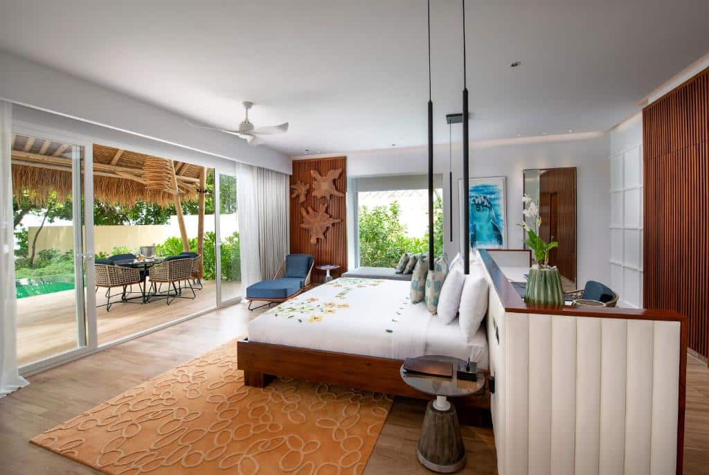 quarto do Emerald Maldives Resort & Spa-Deluxe All Inclusive, com cama enorme, balcão e poltronas com janela grande de vidro e porta também, com varanda com piscina, mesinhas e cadeiras