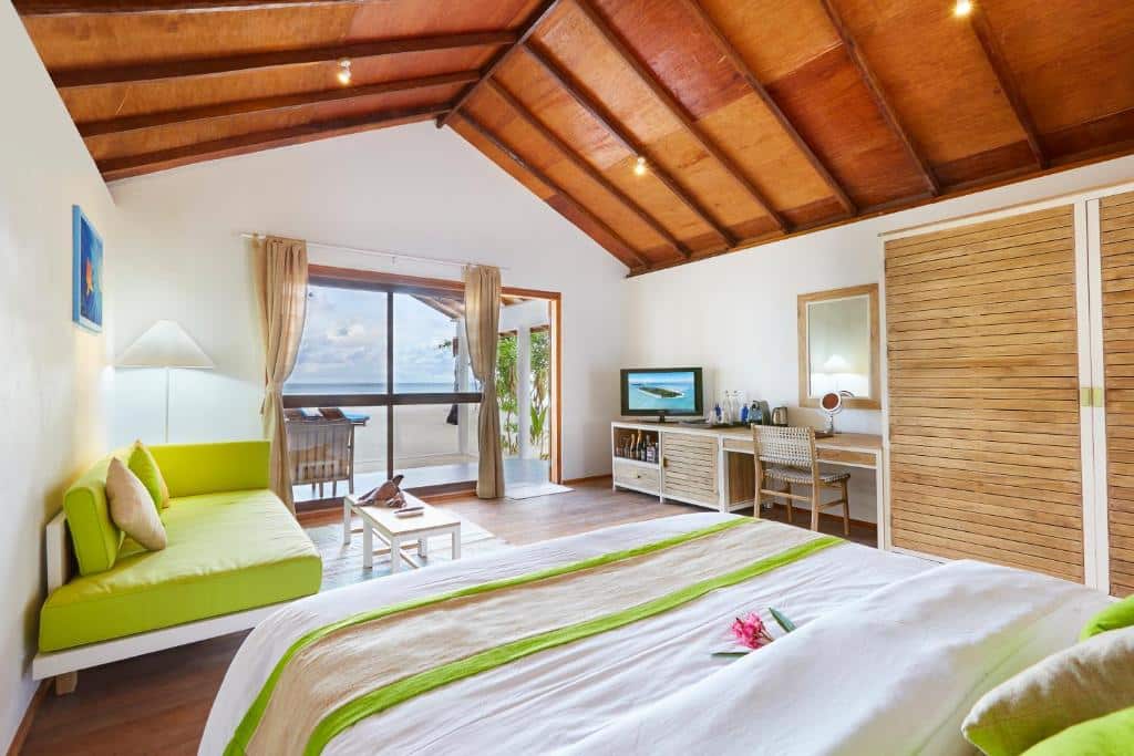 quarto do Innahura Maldives Resort, com cama grande, guarda-roupa de madeira, sofá, tv e outros móveis com porta de vidro que dá para varanda com vista do mar