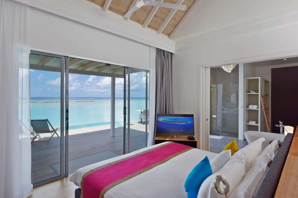 quarto do Kuramathi Maldives com cama de casal grande, tv e banheiro com porta de vidro grande com deck que dá acesso à praia