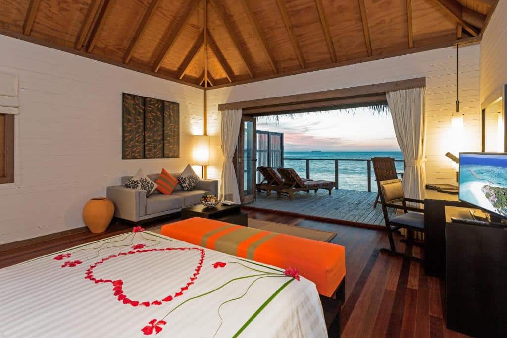 quarto do Meeru Island Resort & Spa, com cama de casal grande com um coração desenhado em pétalas vermelhas, com sofá, janela de madeira, cadeira, mesa e uma varanda com vista pro mar