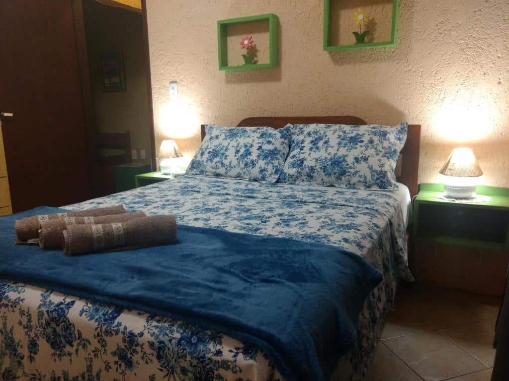 quarto da Pousada Santa Genoveva, com cama de casal em detalhes floridos azuis, quadros com ninhos, luzes de ambos os lados da cama com mesinhas de cabeceira e guarda-roupa também de madeira