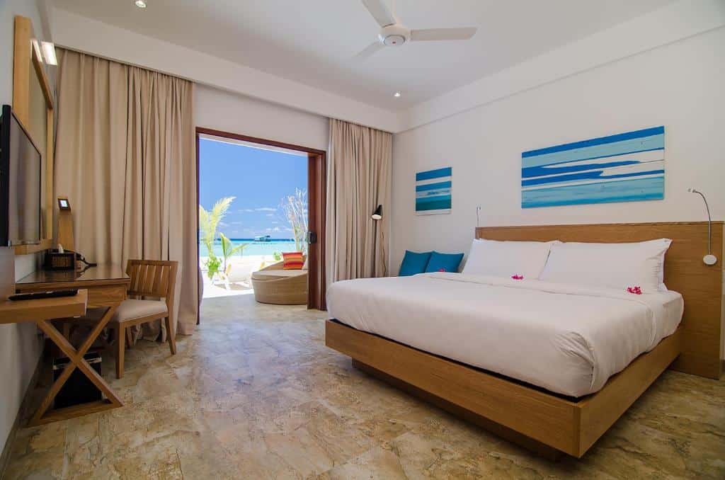 quarto do Summer Island Maldives Resort, um dos resorts nas Maldivas, com cama de casal bem grande, quadros azuis na parede, mesa, cadeiras e uma porta grande para a varanda com poltronas e vista para a praia