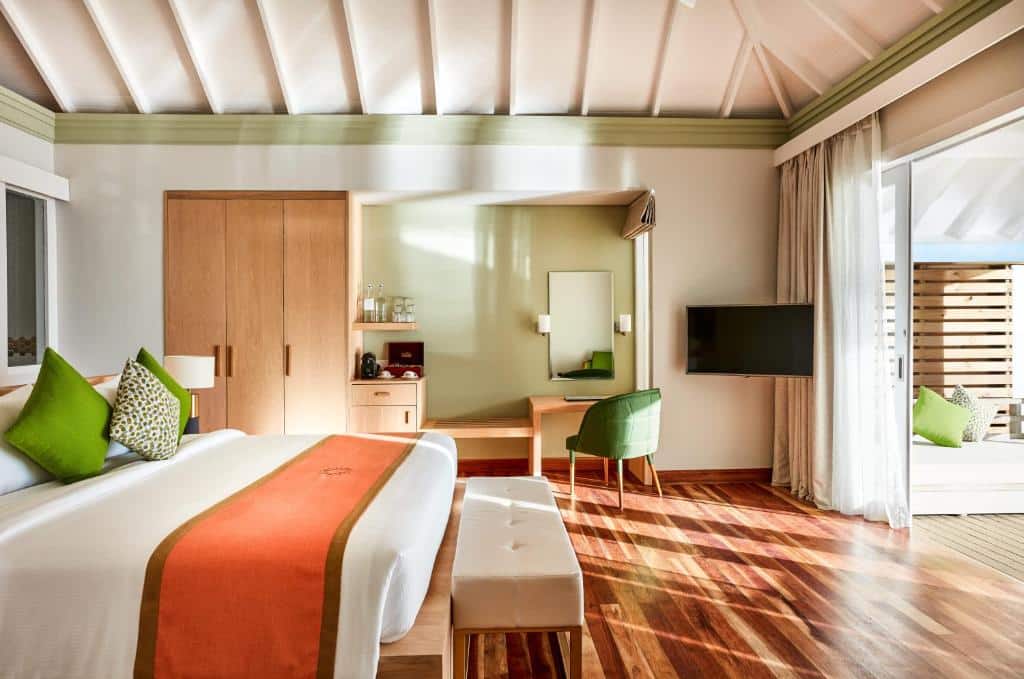quarto do Sun Siyam Olhuveli, com cama grande de casal, guarda-roupa, cadeira com mesa, tv e porta grande que conecta a uma varanda tudo em tons de laranja e verde