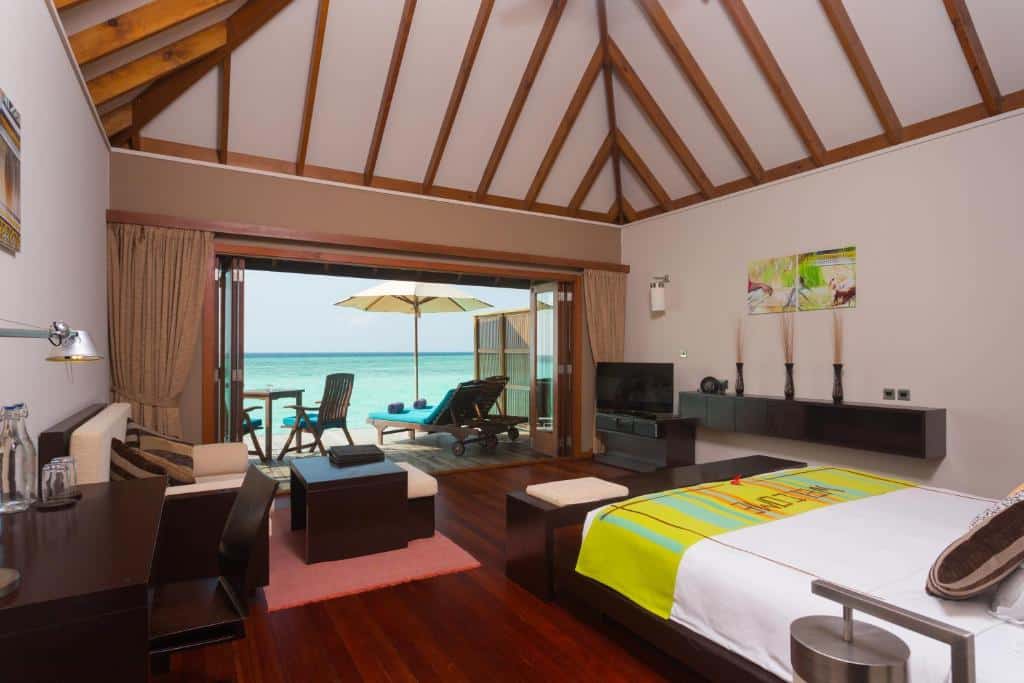 quarto do Veligandu Island Resort & Spa, com cama grande de casal, sofá cama, mesinha e cadeiras, tv e porta grande para a varanda com vista pro mar