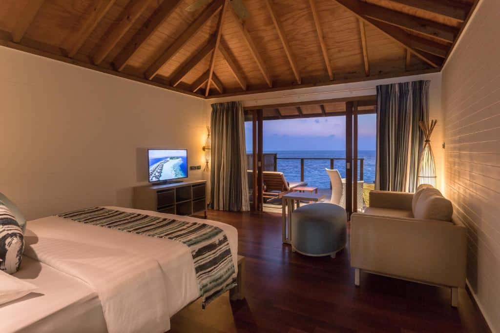 quarto com cama de casal sofá e mesinha de centro em frente a uma televisão e varanda com cadeiras em frente ao mar no Vilamendhoo Island Resort & Spa