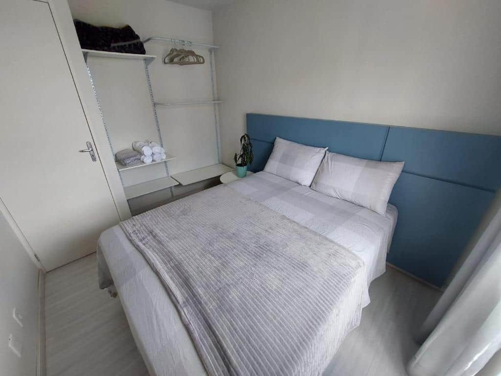quarto do Apartamento Próximo ao Aeroporto, em Curitiba, com cama de casal com roupas de cama brancas e cinza claras, cabeceira azul clara e armários e guarda-roupa abertos, com uma mesinha ao lado da cama