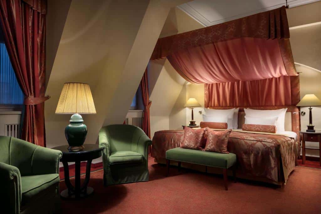 Quarto do Art Nouveau Palace Hotel com uma cama de casal, duas poltronas e uma pequena mesinha oval com um abajur, carpete vermelho e duas janelas com cortinas