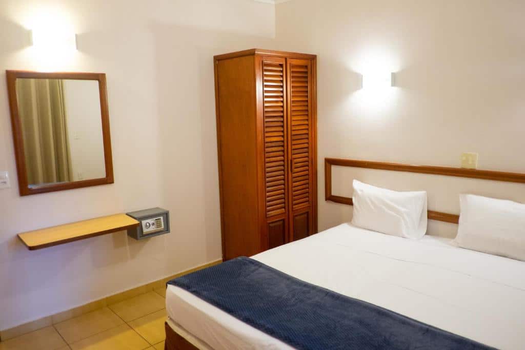 Quarto do Atibaia Residence Hotel & Resort com cama de casal do lado direito e do lado esquerdo um pequeno guarda-roupa ao lado de um cofre e um espelho.