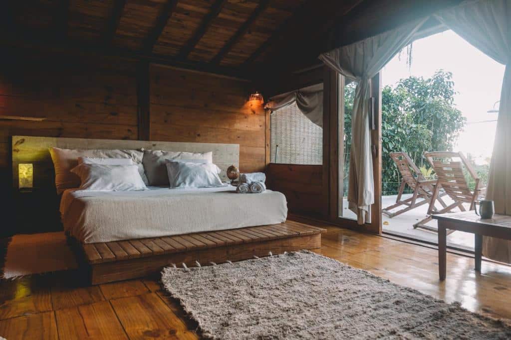 Quarto do Casa Hotel Ocean com cama de casal grande do lado esquerdo portas de madeiras amplas com acesso a varanda com cadeiras de madeiras. Representa airbnb em Trancoso.