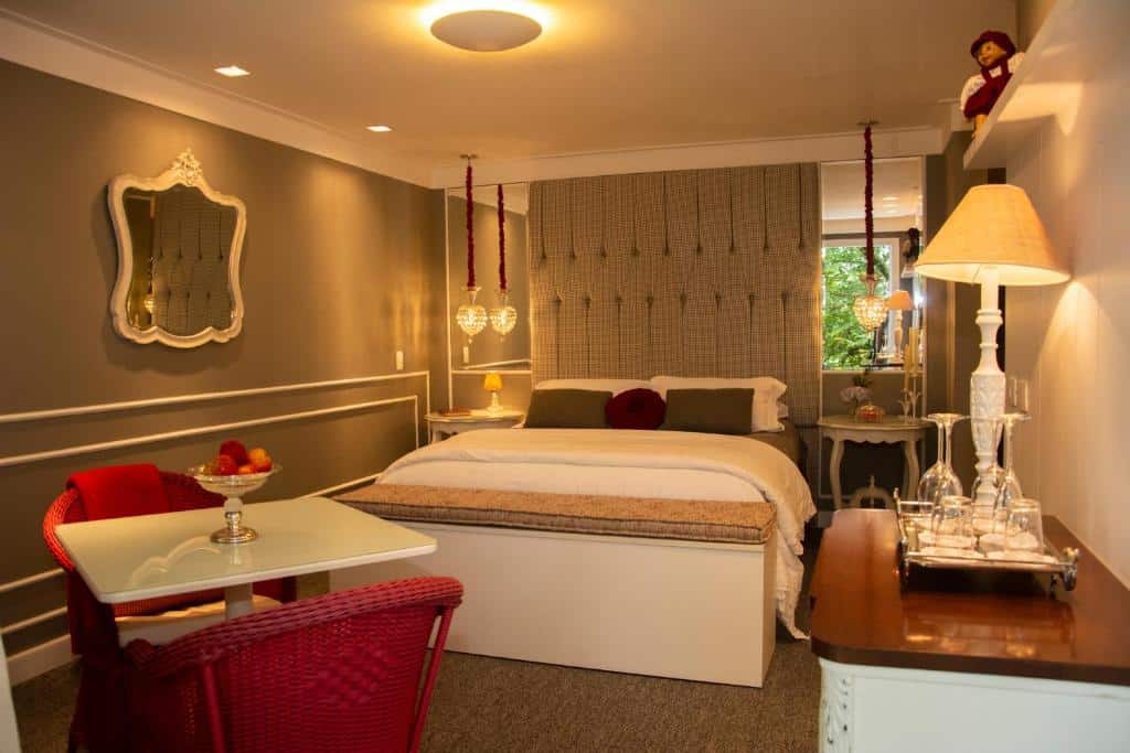 Quarto luxuoso do Casa Regaleira Hotel Boutique com uma cama de casal, diversas iluminações com abajures e candelabros pendurados nas laterais da cama, uma mesinha com duas cadeiras vermelhas, chão de carpete, uma cômoda de madeira com taças e um abajur sob o móvel