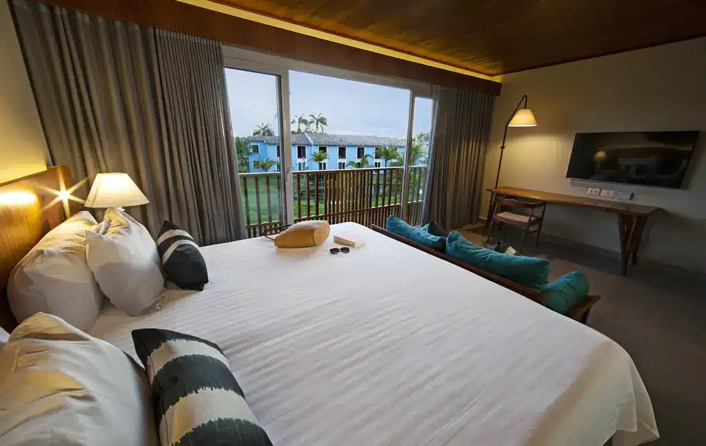 Quarto do Club Med Trancoso com cama de casal, luminária do lado esquerdo da cama, em frente a cama um pequeno sofá com almofadas verdes que tem à frente uma mesa de trabalho de madeira com TV presa na parede.
