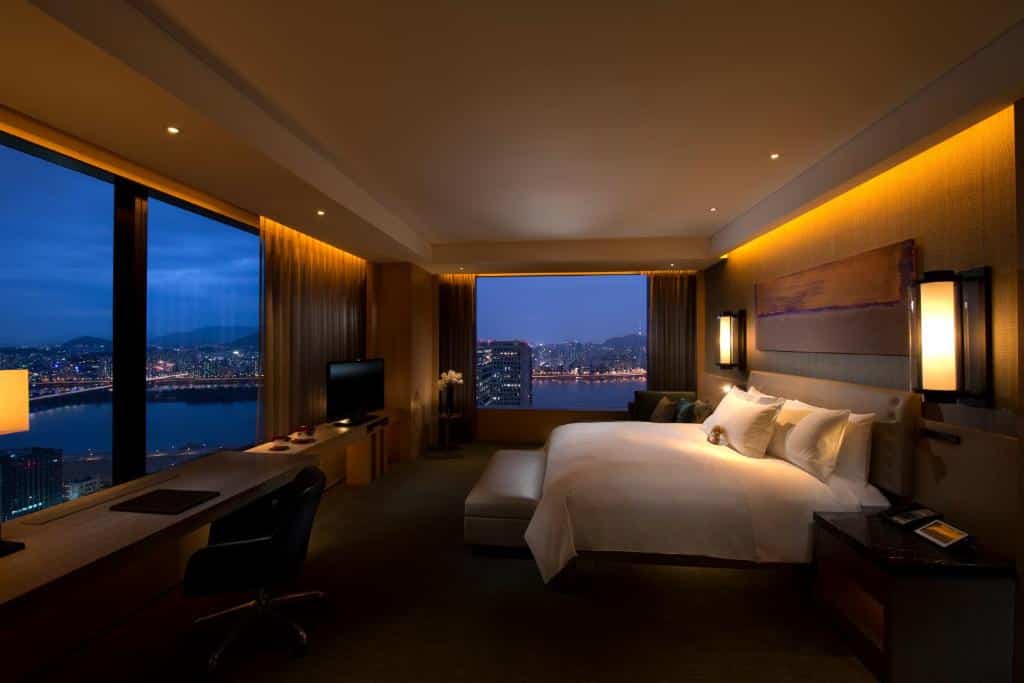 Quarto do Conrad Seoul com janelas panorâmicas, uma bancada com uma televisão e espaço para um notebook, cama de casal, uma poltrona e duas luminárias