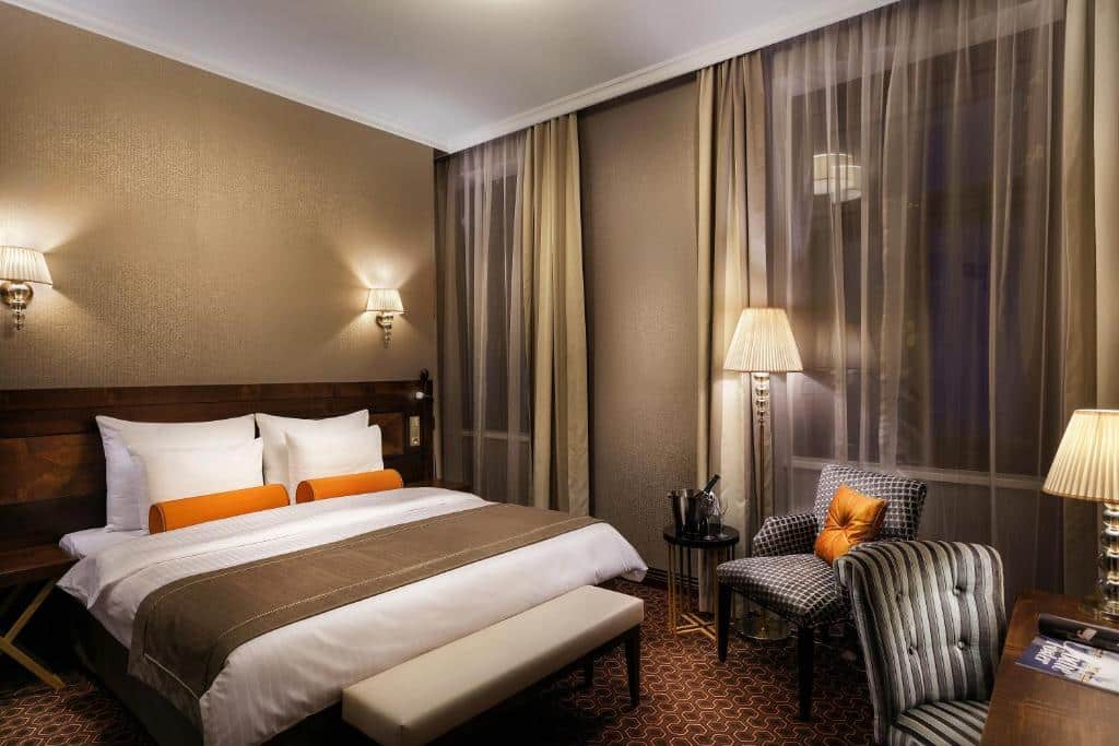 Quarto do COSMOPOLITAN Hotel Prague com duas janelas com cortinas, uma cama de casal, carpete marrom, um abajur de chão, uma mesa de trabalho com uma cadeira